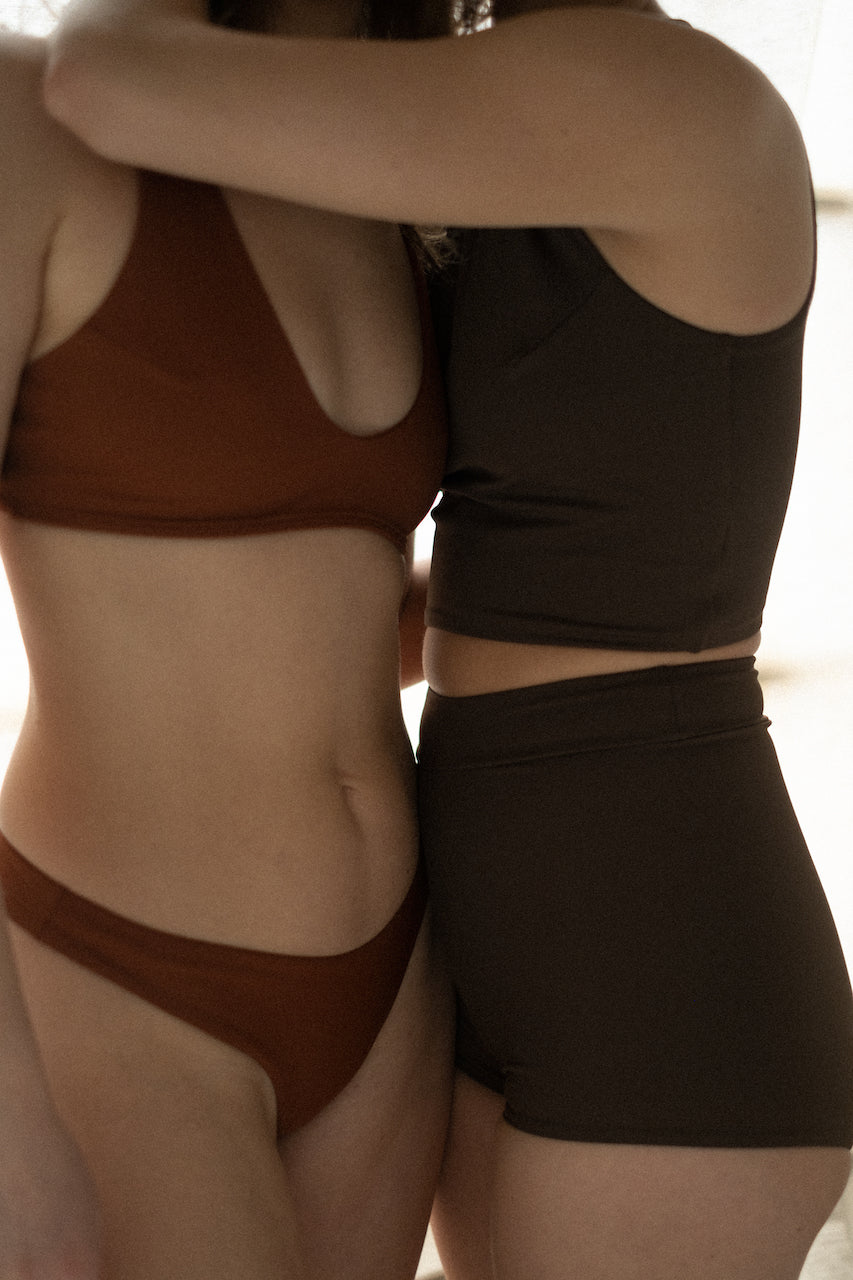 Calm in every transition—two women in Tumla’s true black and burnt orange swimwear enjoy a peaceful yoga routine by the water