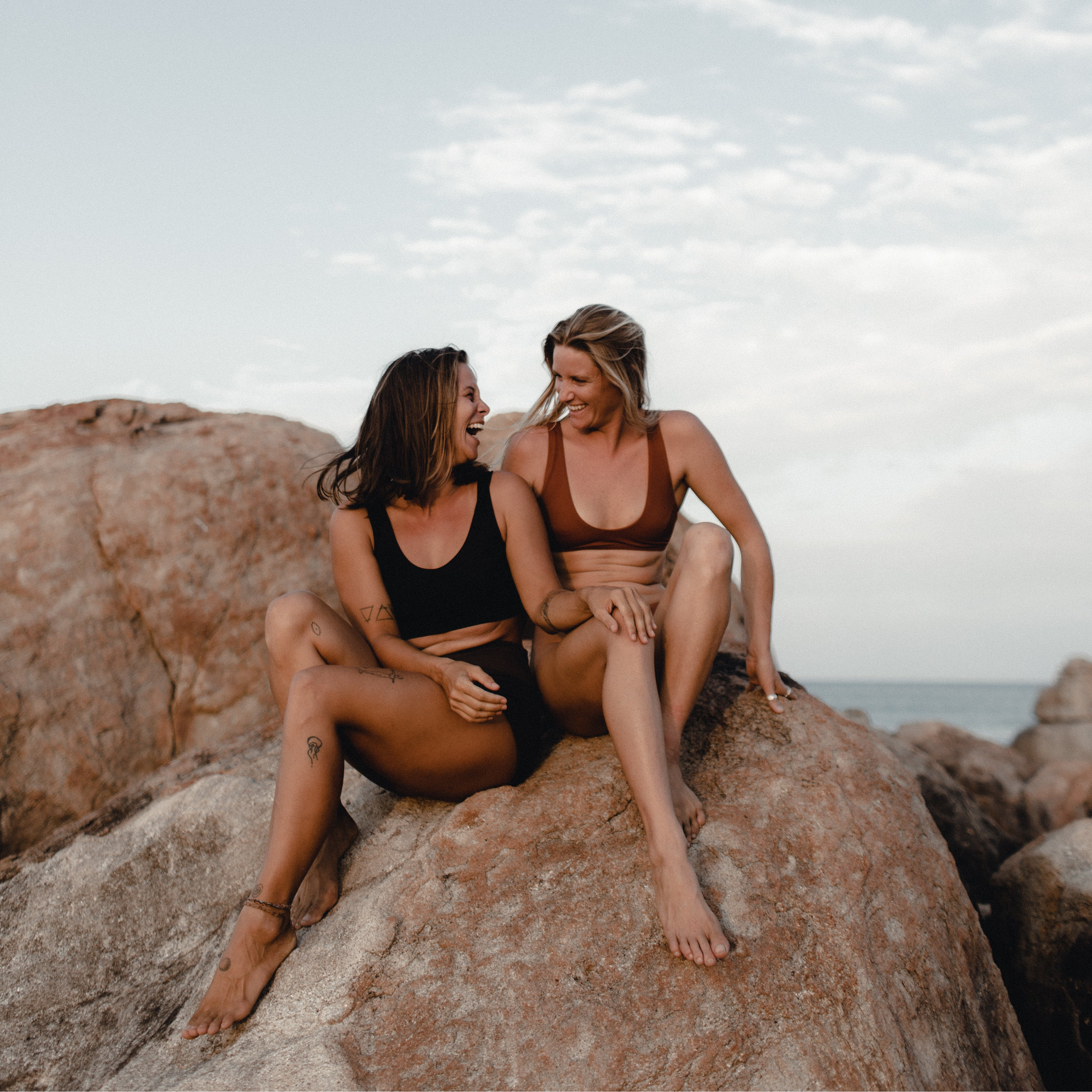 Two women wearing Tumla’s high-performance swimwear sit on rocky shores, showcasing confidence, strength, and adventure-ready design