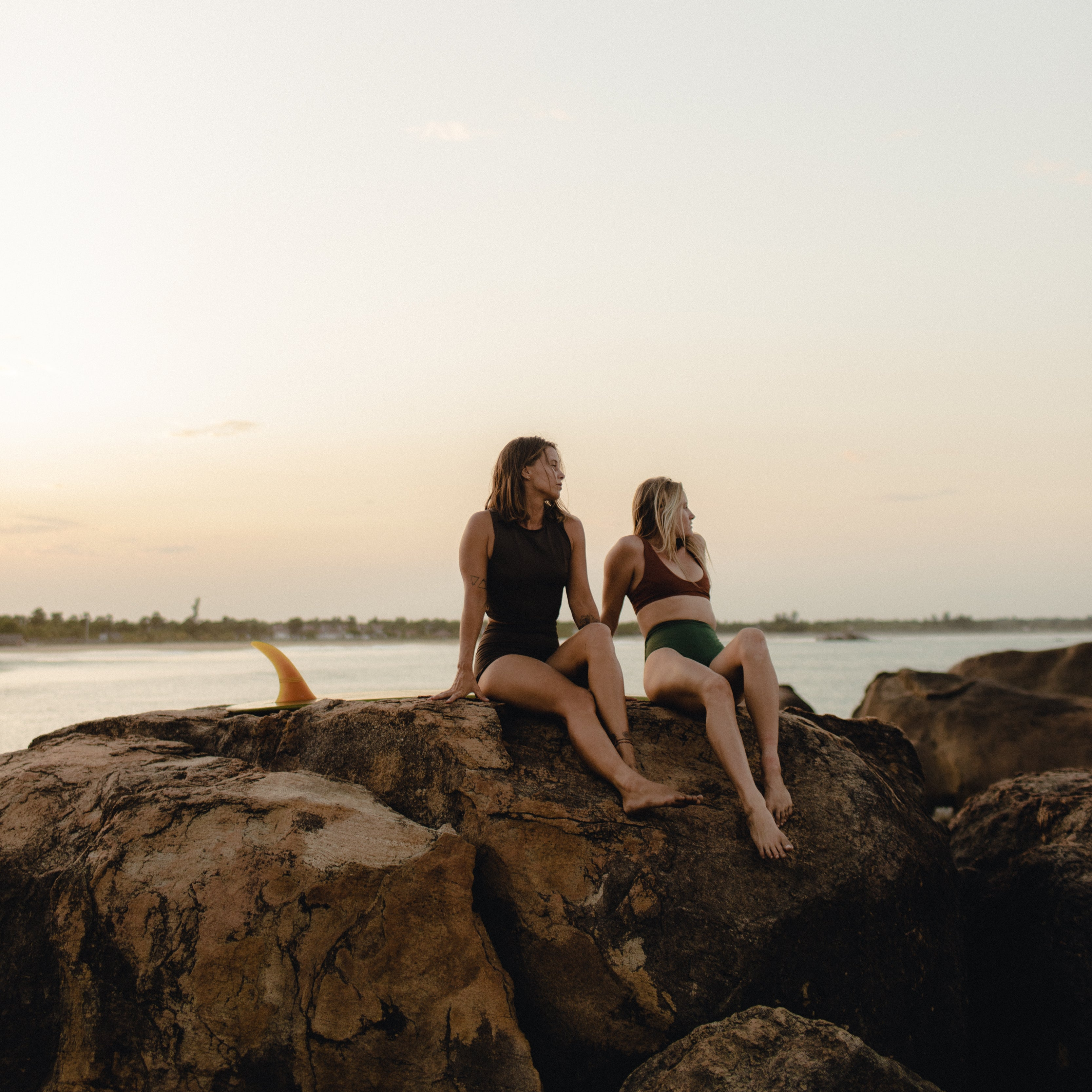 Two women wearing Tumla functional swimwear sit on coastal rocks at sunset, embracing nature and adventure. Designed by women, for women, Tumla bikinis offer comfort, style, and performance for an active lifestyle in and out of the water