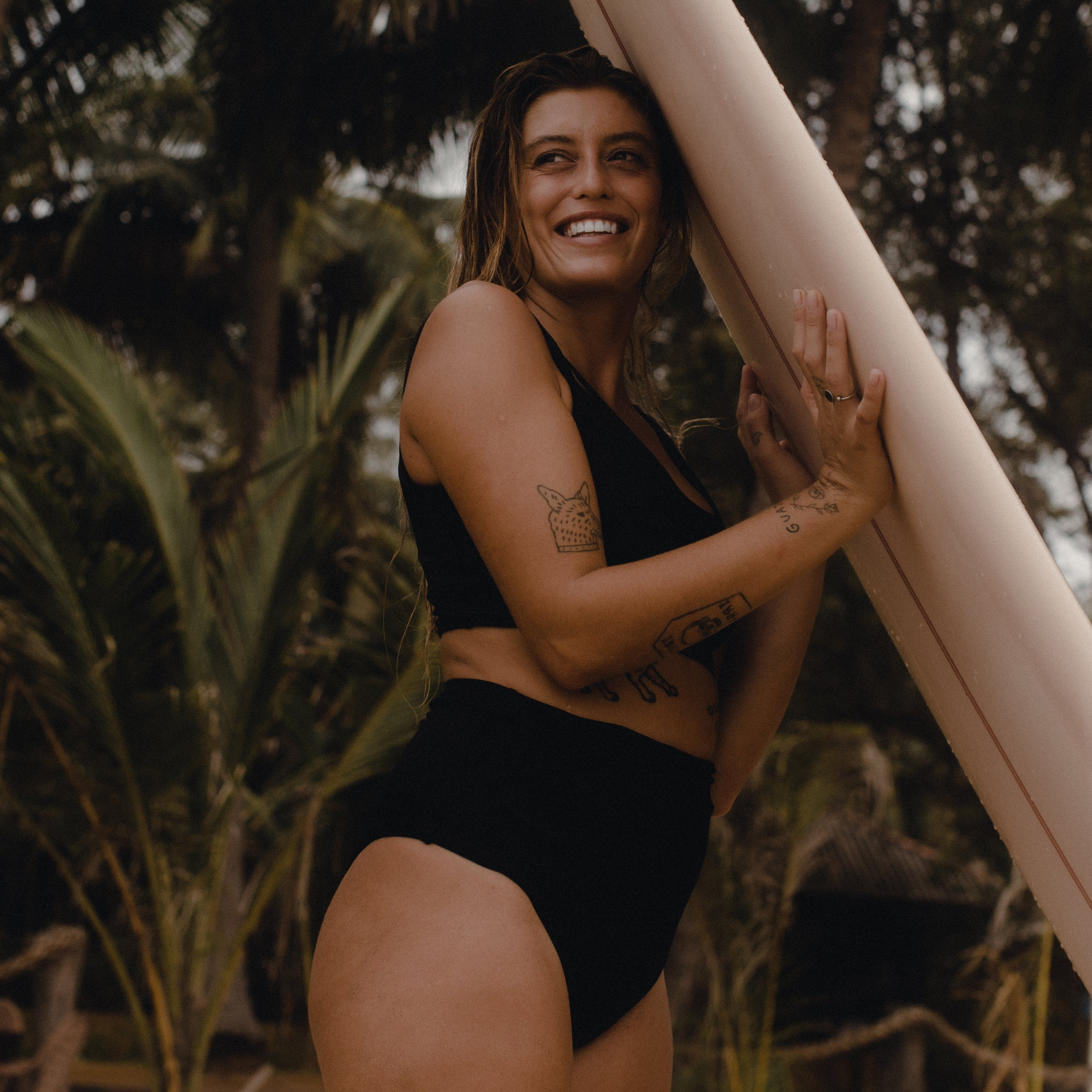 Woman in a black bikini holding a surfboard with tropical plants in the background